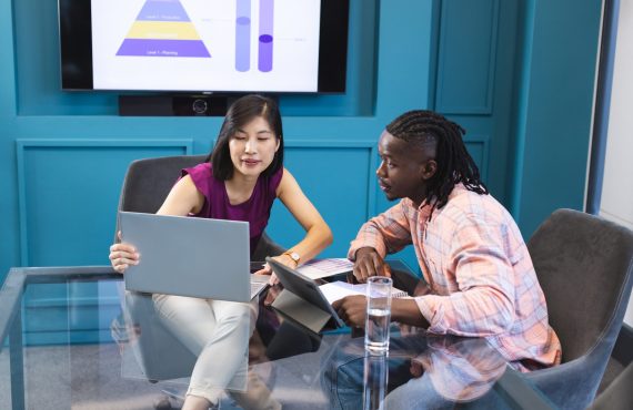 Asian woman and African American man looking at laptop screen together in a modern business office. She has black hair, he wears a checkered shirt, both young and focused, unaltered.