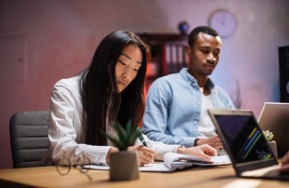 Asian woman and african american man sitting together at modern office and working on tablet and laptop. Competent brokers analyzing charts on trade market during evening time.