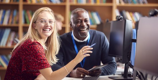 Female University Or College Student Working At Computer In Library Being Helped By Tutor