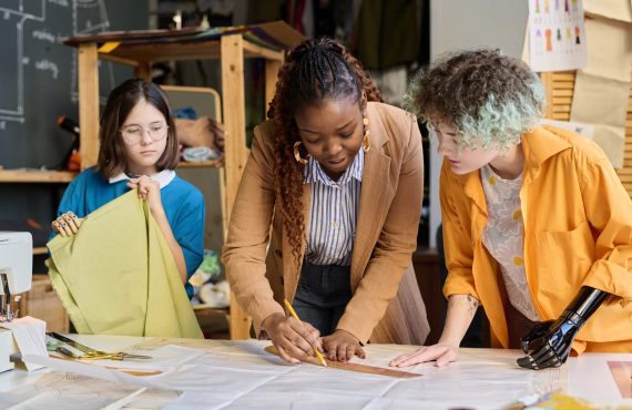 Portrait of Black young woman teaching two girls with disabilities sewing clothing in inclusive atelier studio