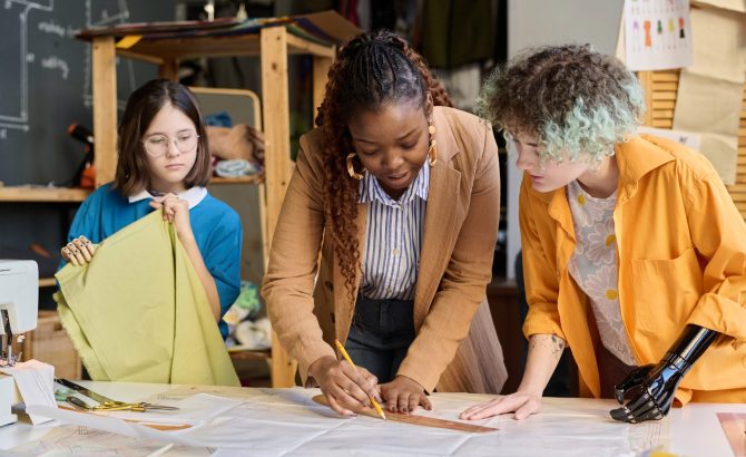 Portrait of Black young woman teaching two girls with disabilities sewing clothing in inclusive atelier studio